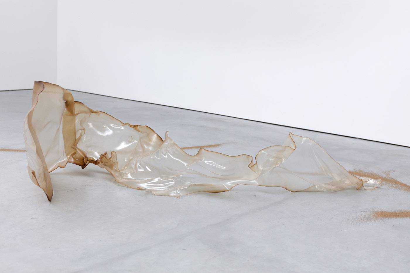 A translucent, skin-like sculptural object made from bound mealworm frass and bio-based plastic lies sprawled across a white gallery floor. The material appears organic and fragile, resembling a shed skin or deflated form. GEOBRIS Polyform (Zustand III), 2025, developed in collaboration with MERGE Cluster, TU Chemnitz.