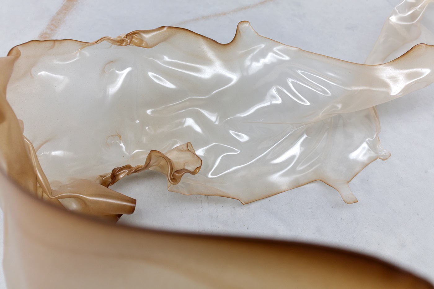 A translucent, skin-like sculptural object made from bound mealworm frass and bio-based plastic lies sprawled across a white gallery floor. The material appears organic and fragile, resembling a shed skin or deflated form. GEOBRIS Polyform (Zustand III), 2025, developed in collaboration with MERGE Cluster, TU Chemnitz.